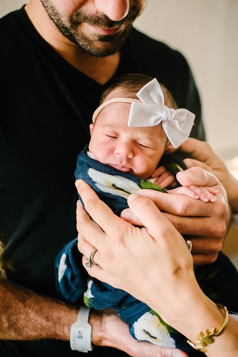 baby girl in navy blue swaddle and white bow is held by her parents during a hospital newborn session
