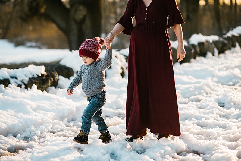 mother and toddler son hold hands and walk through snow during family maternity session