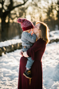 pregnant mom wearing maroon dress holds toddler sun during golden hour snowy session