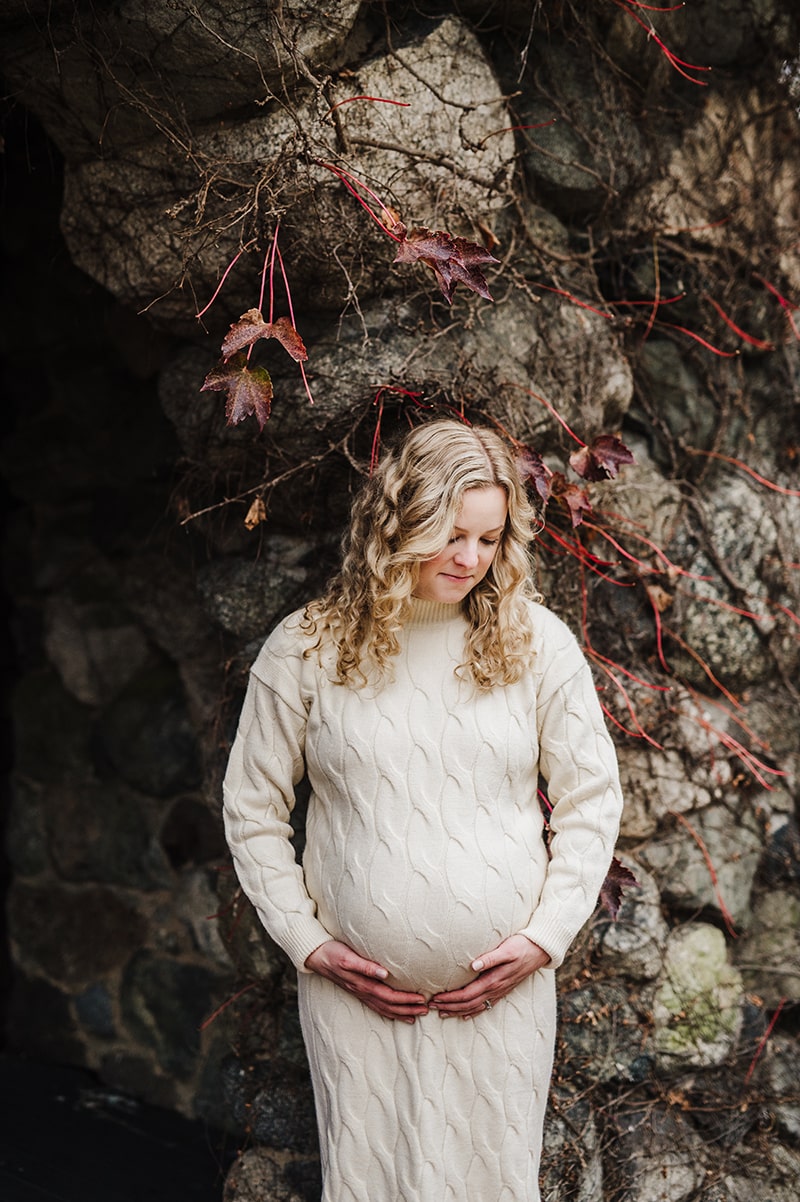 mom cradles her baby bump in front of a stone walk covered in red ivy during her maternity session