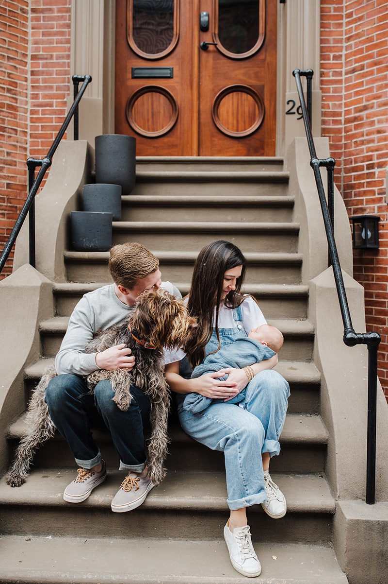 family of 3 snuggles on their front stoop in Boston with their newborn baby and pup