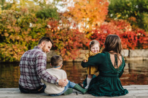 family of four sits on a dock enjoying the Charles River and the beautiful fall foliage in Boston