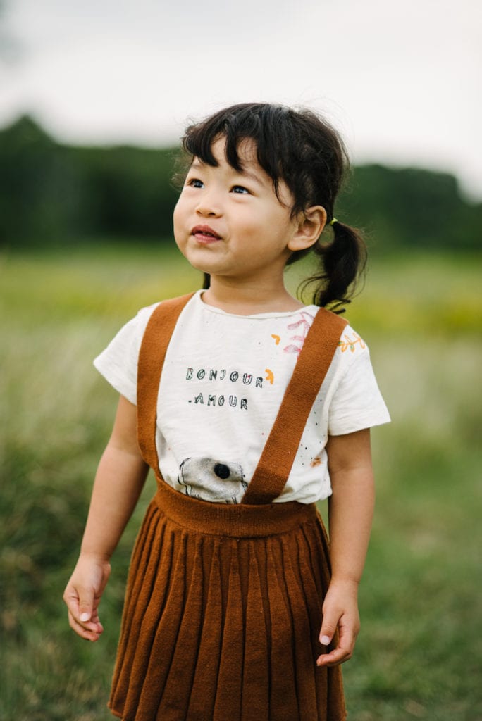 Family Photographer, a young girl stands in a grassy field in her overalls happy
