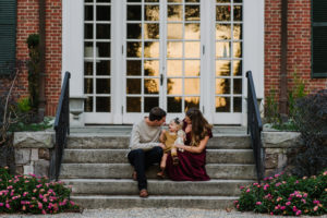 Family Photographer, a mom and dad sit on the steps outside their home with their baby daughter