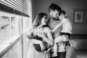 black and white image of family of four standing by a window snuggling their new addition