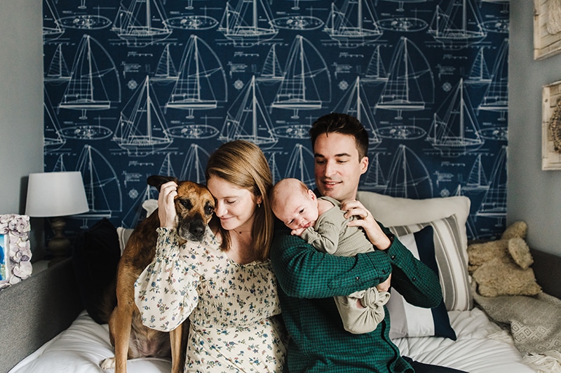 new family of 3 snuggles with their newborn son and dog in front of beautiful navy blue nautical accent wall