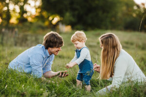 family of 3 plays in the grass with toddler son as he pulls grass from the field