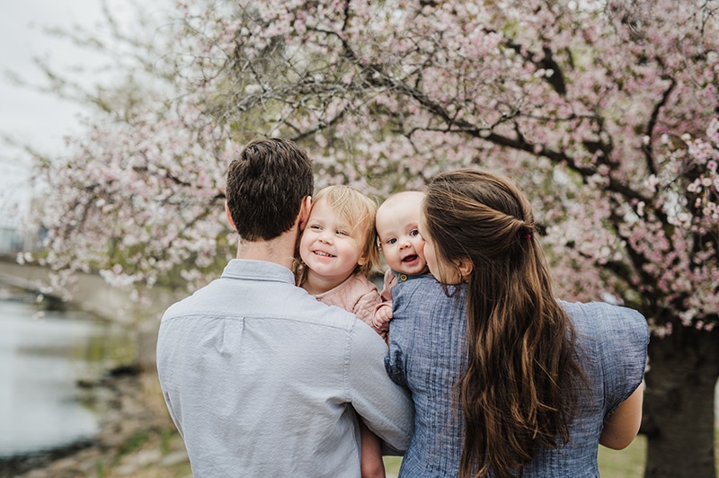 family of four snuggling in front of a cherry tree while holding their young girls up over their shoulders
