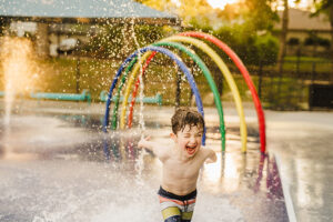 pre-school aged boy runs excitedly through a rainbow splash pad during golden hour